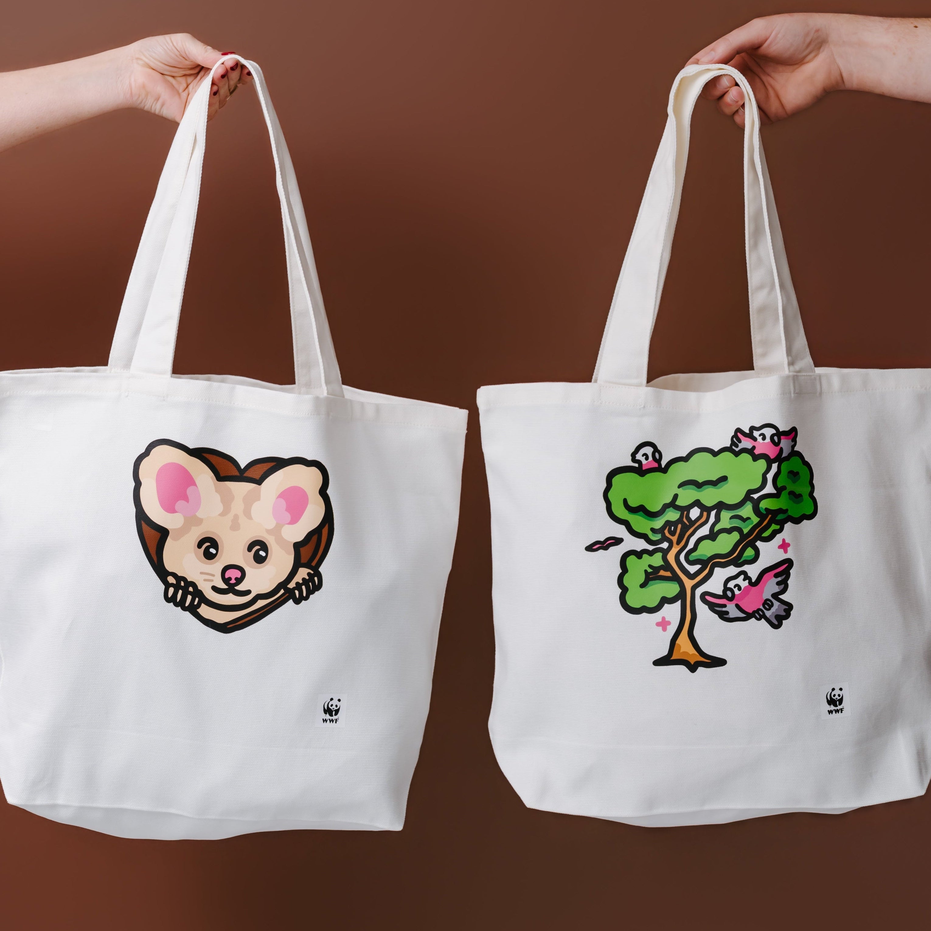 Two white canvas tote bags held up by human arms against a solid brown background. The left tote bag features a cute greater glider illustration, and the right tote bag features a colorful cartoon tree with a pink flower.