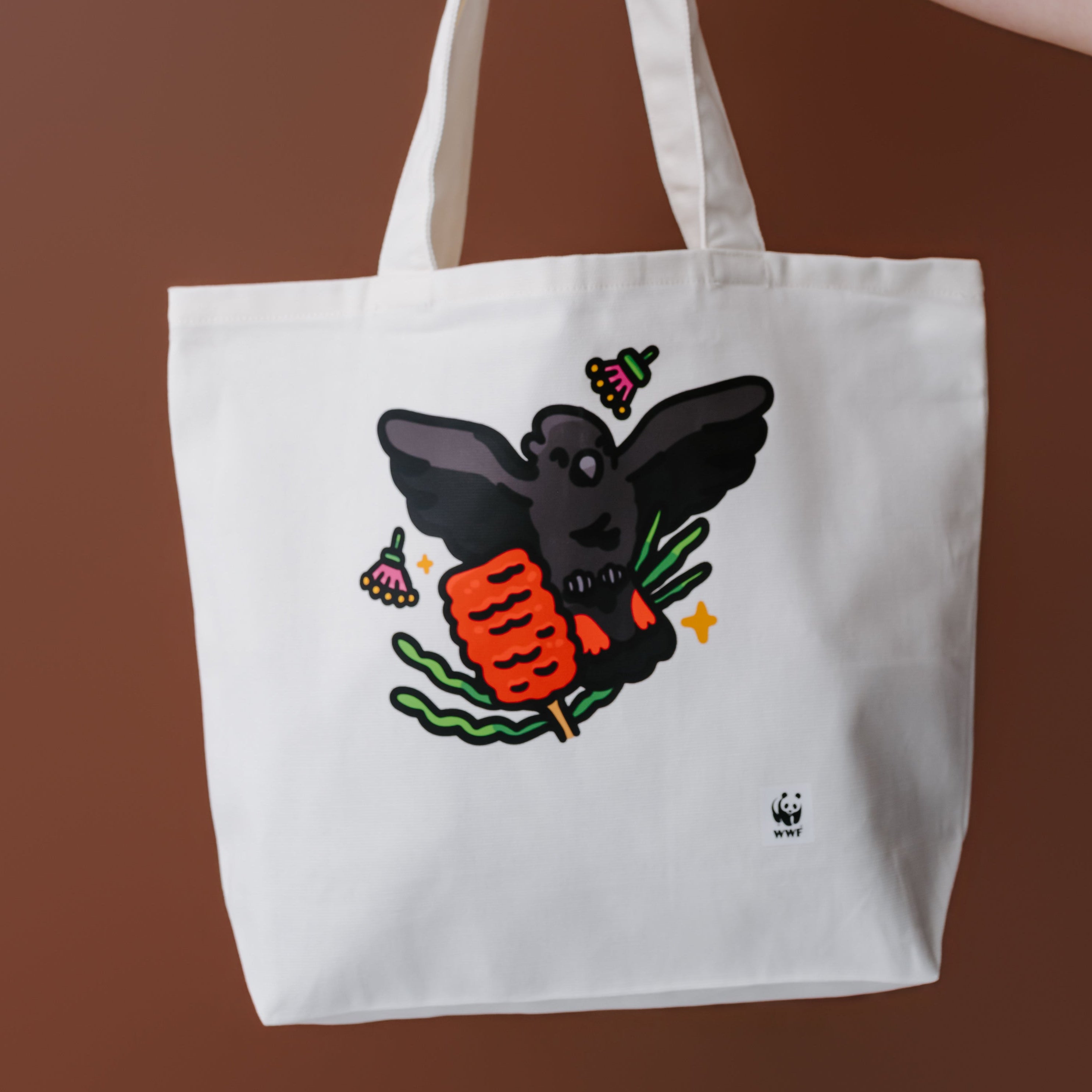A white canvas tote bag with a graphic of a glossy black cockatoo, held by a person's tattooed arm against a solid brown background. 