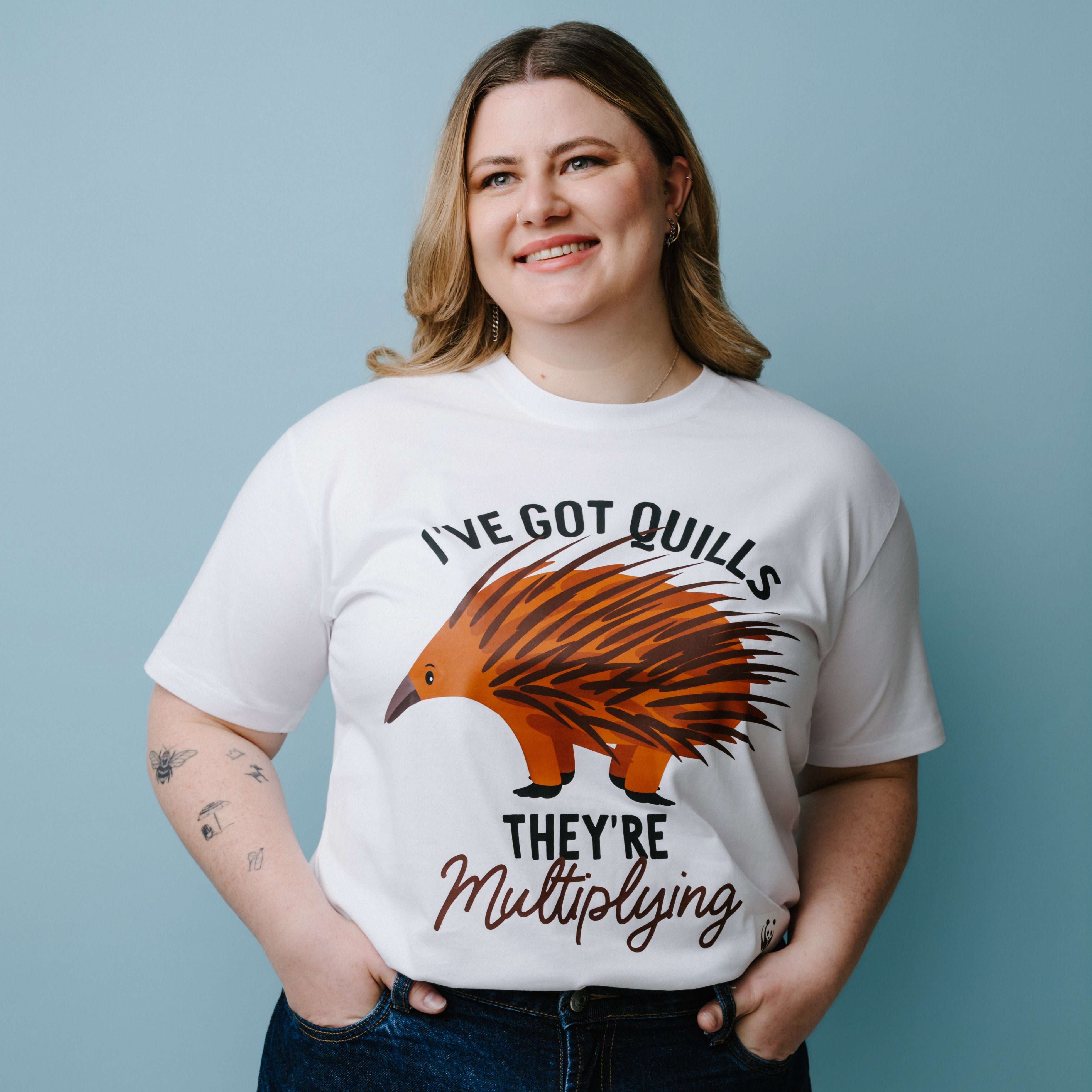 Person wearing a white t-shirt with a graphic of an echidna and text, "I've Got Quills They're Multiplying" against a blue background. The product is a WWF-Australia T-Shirt.