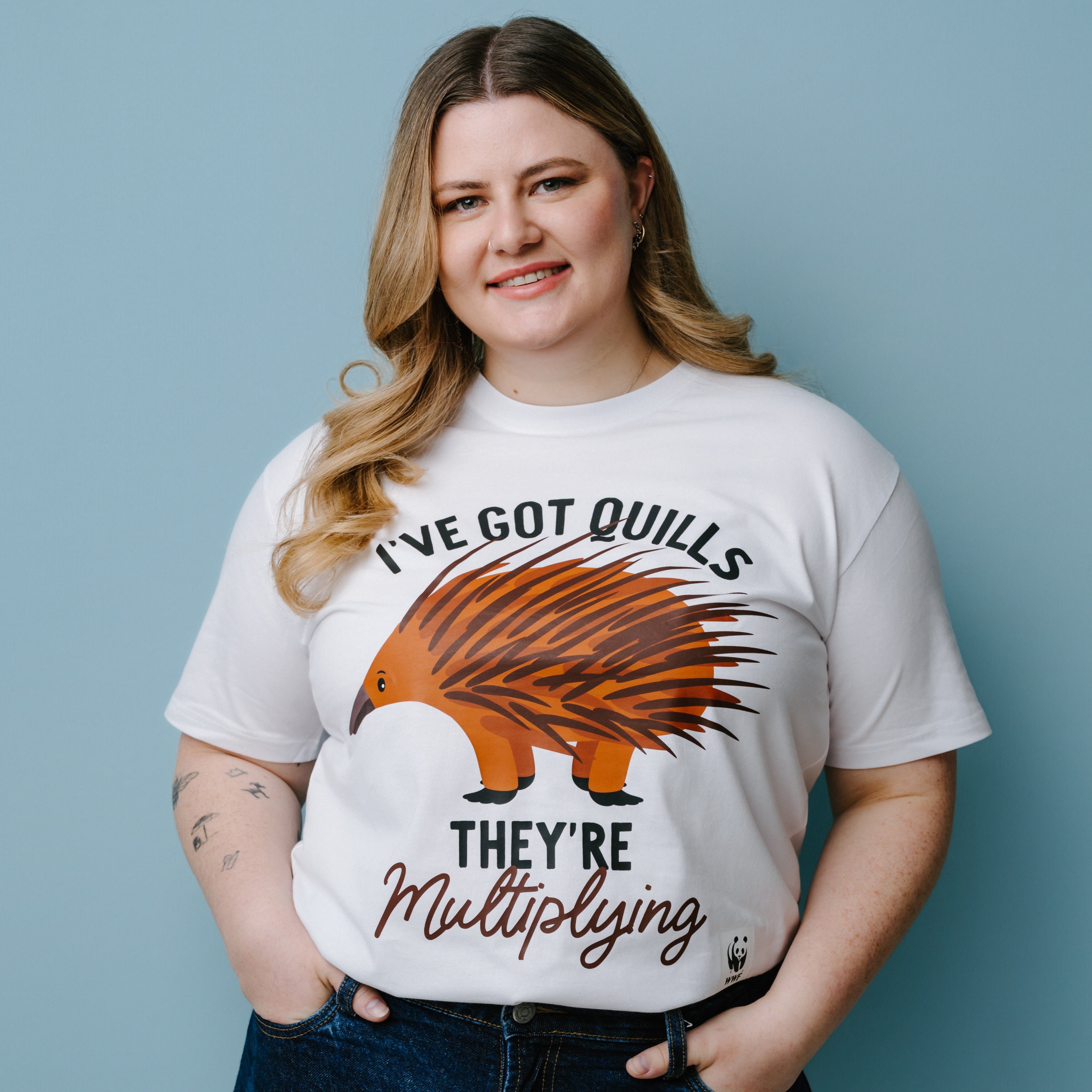 Person wearing a white t-shirt with a graphic of an echidna and text, "I've Got Quills They're Multiplying" against a blue background. The product is a WWF-Australia T-Shirt.
