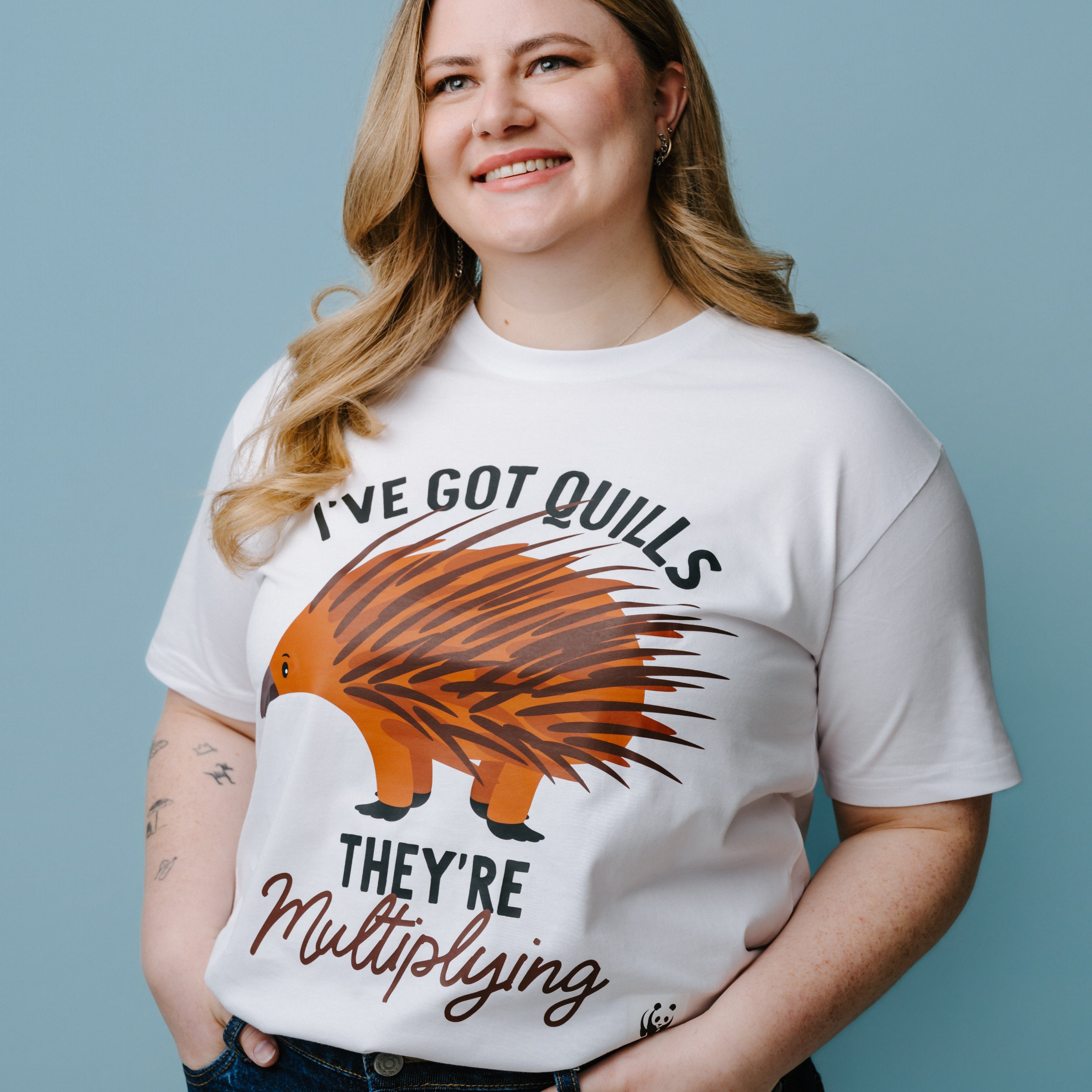 Person wearing a white t-shirt with a graphic of an echidna and text, "I've Got Quills They're Multiplying" against a blue background. The product is a WWF-Australia T-Shirt.