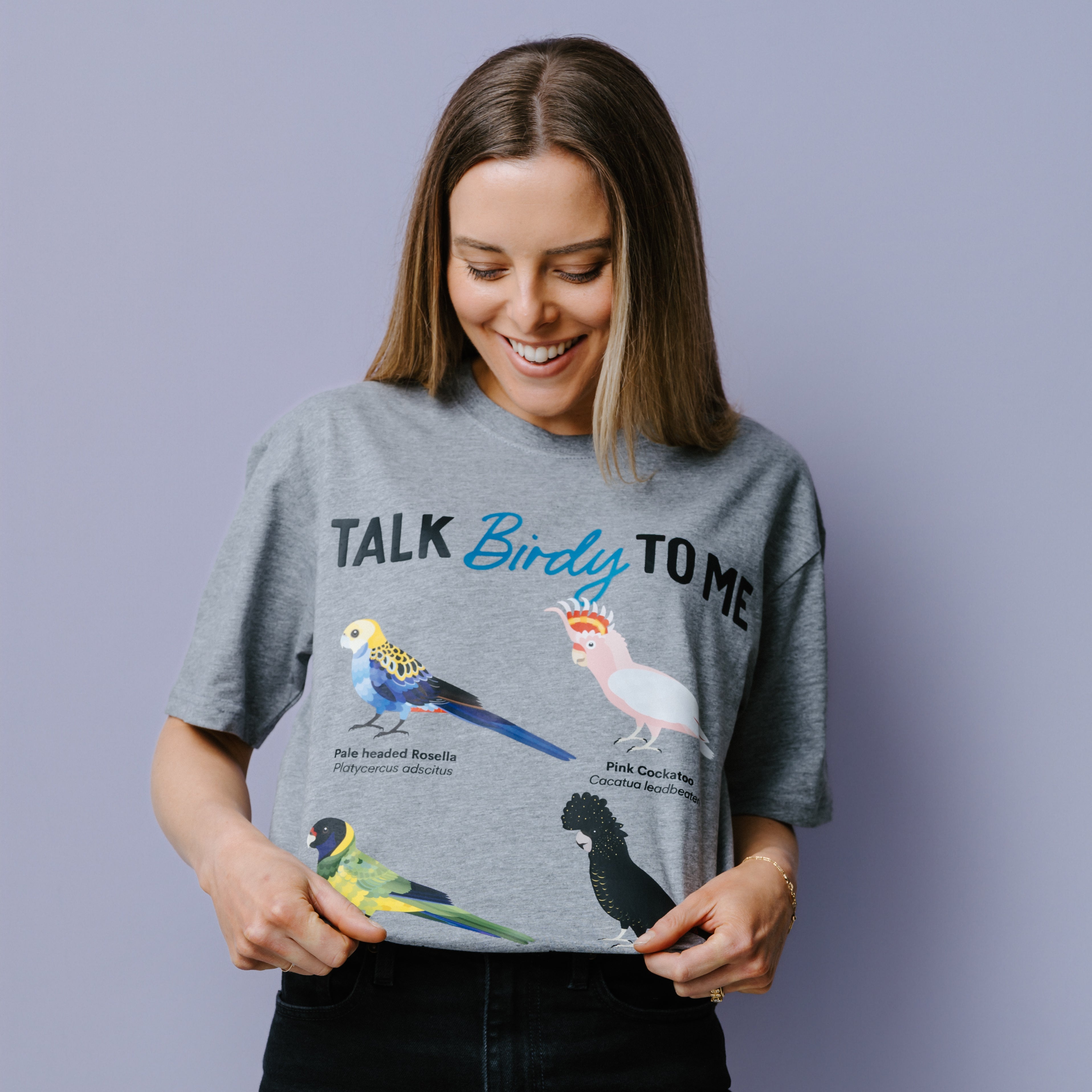 Woman wearing a grey WWF-Australia t-shirt with bird illustrations and text, "Talk Birdy To Me against a purple background.