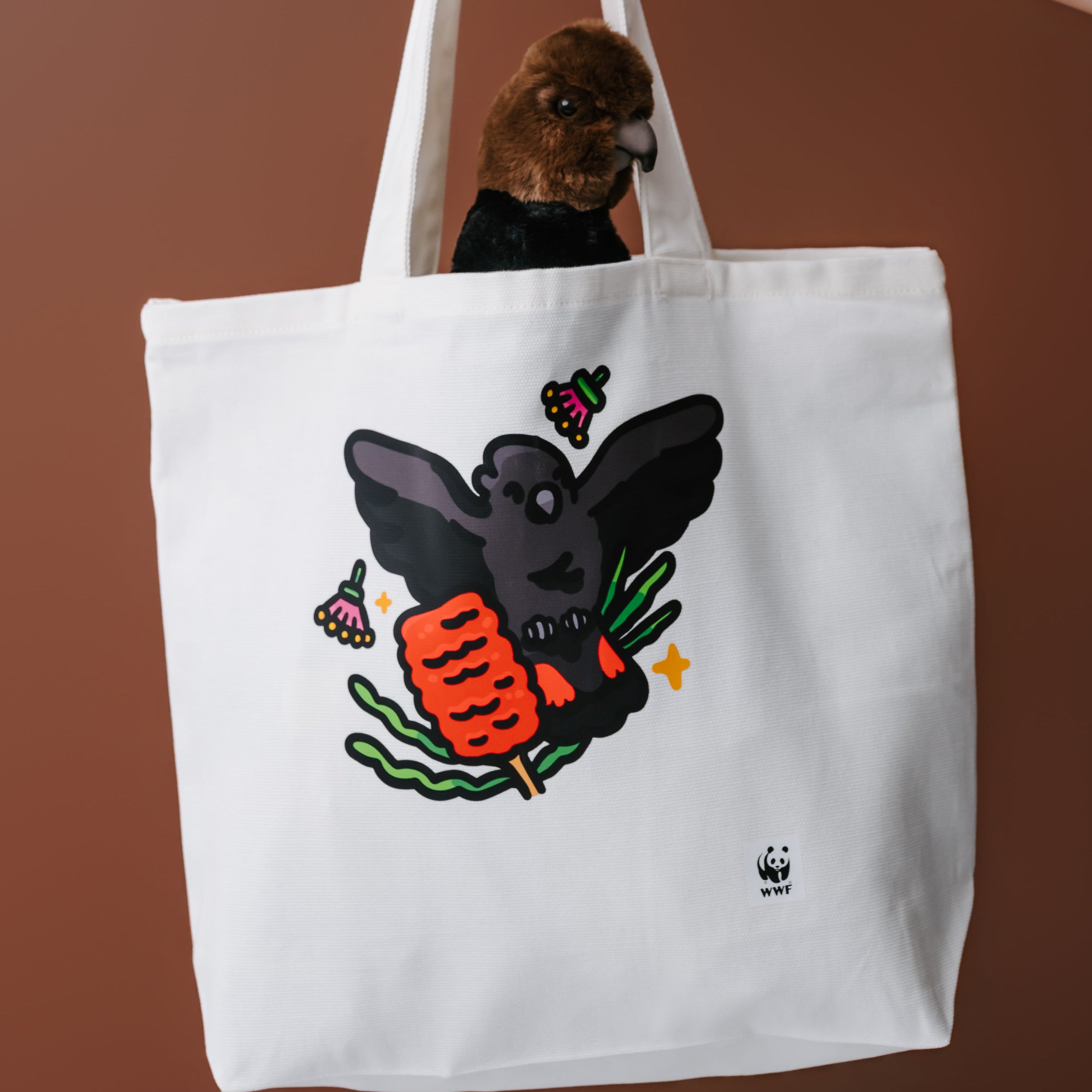 A white canvas tote bag with a graphic of a glossy black cockatoo, held by a person's tattooed arm against a solid brown background. 