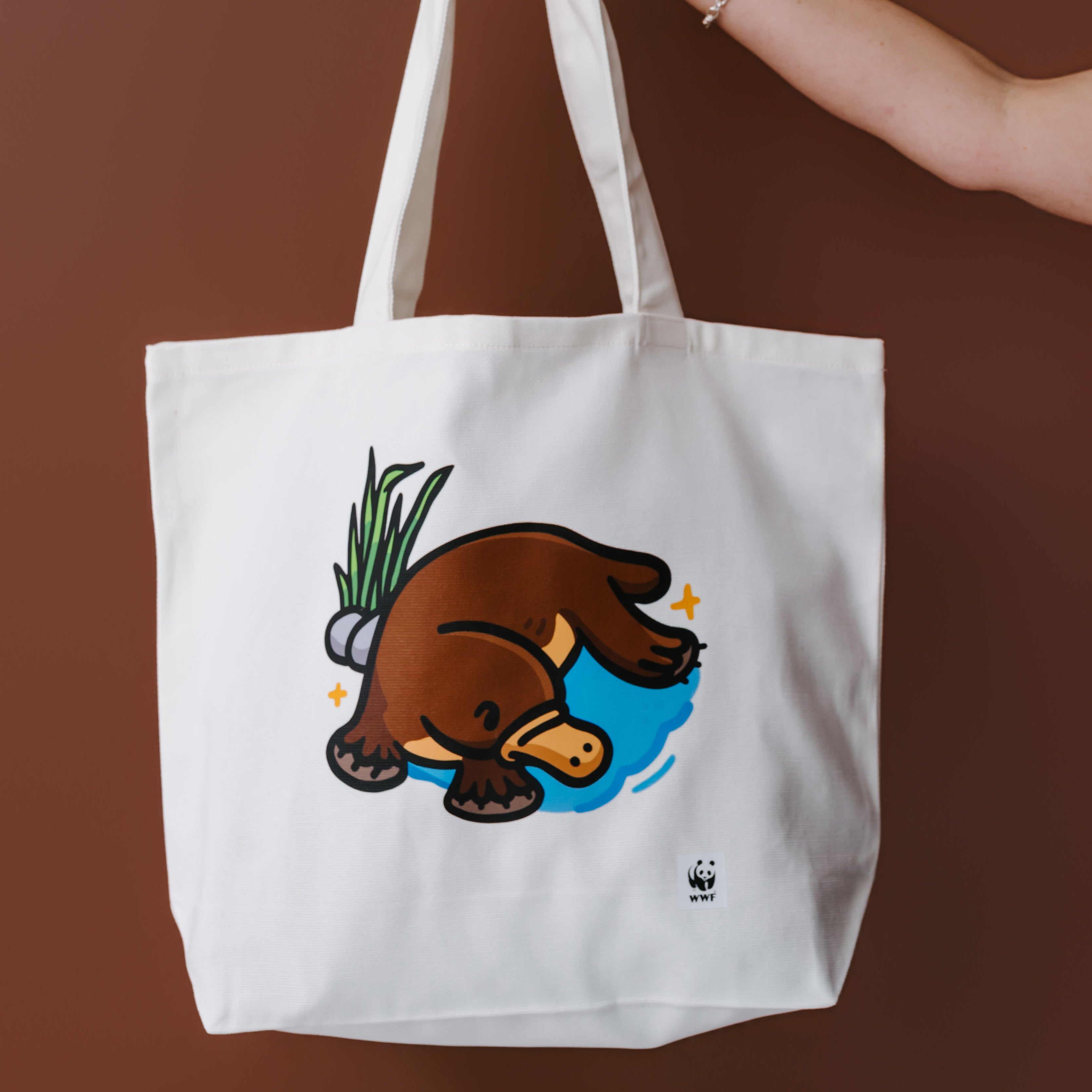A white canvas tote bag with a cartoon platypus illustration, held by a person's hand against a solid brown background.