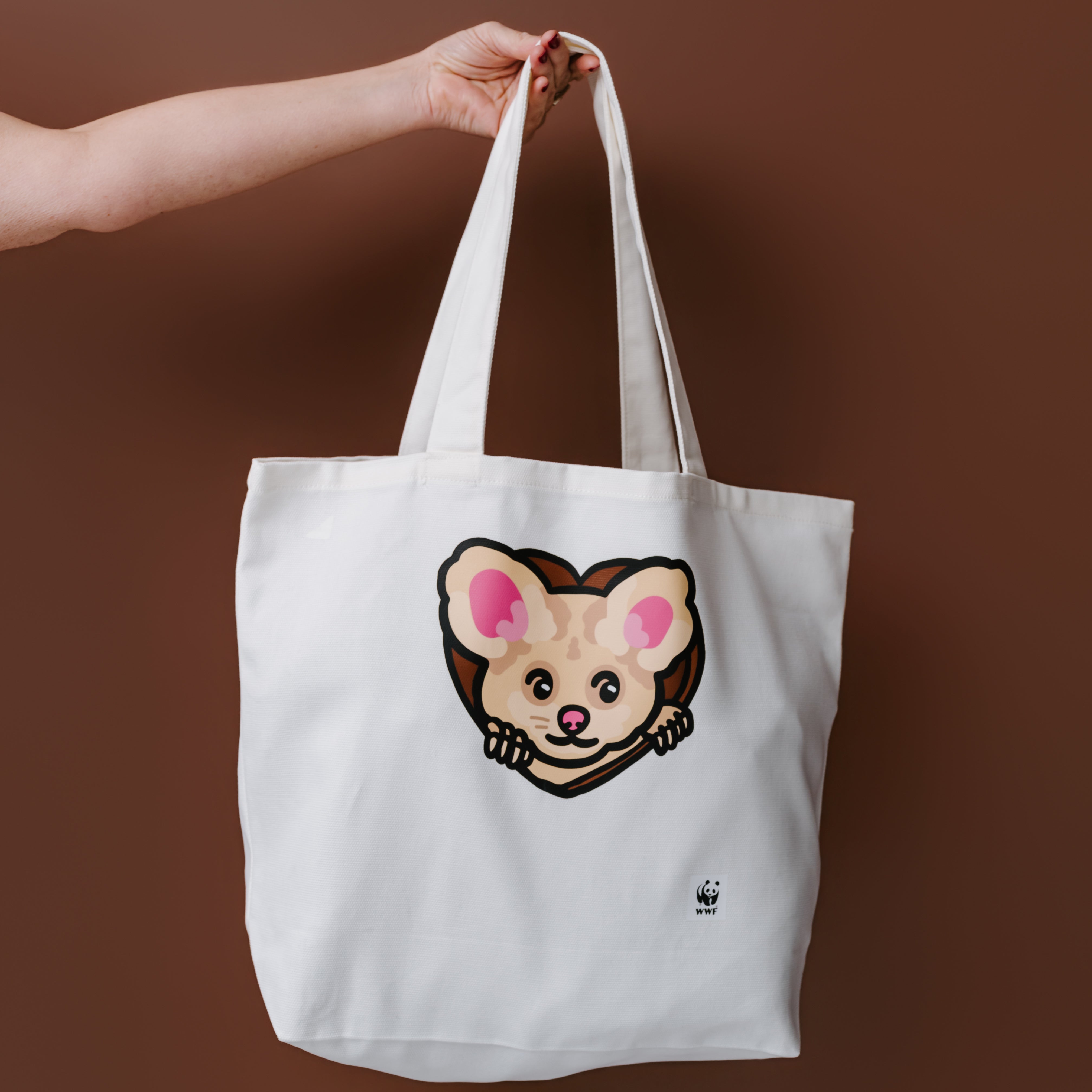 An arm holding up a WWF-Australia Night Puff Greater Glider Tote bag against a brown background