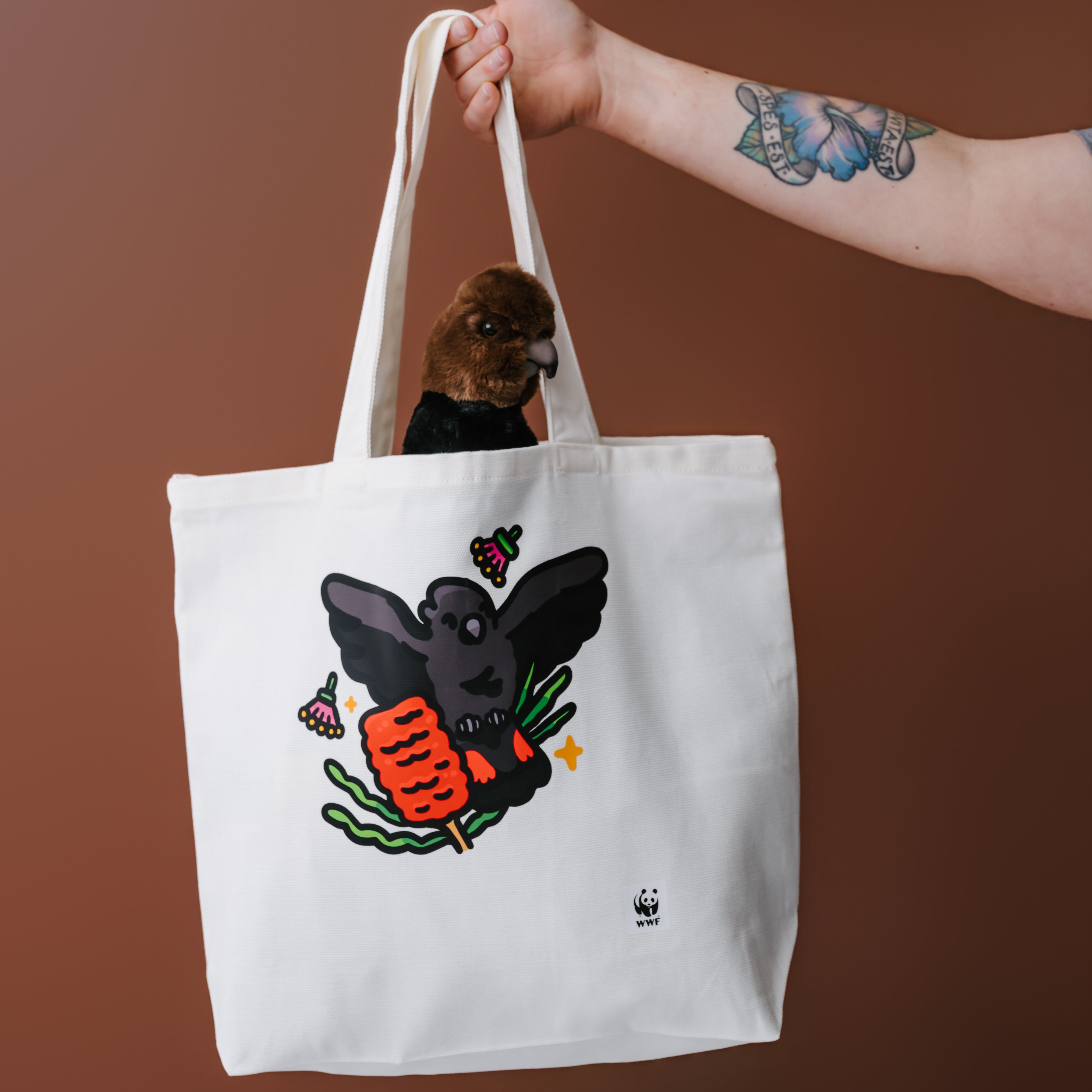 A white canvas tote bag with a graphic of a glossy black cockatoo, held by a person's tattooed arm against a solid brown background. The bag contains a glossy black cockatoo plush sticking out the top of the bag.