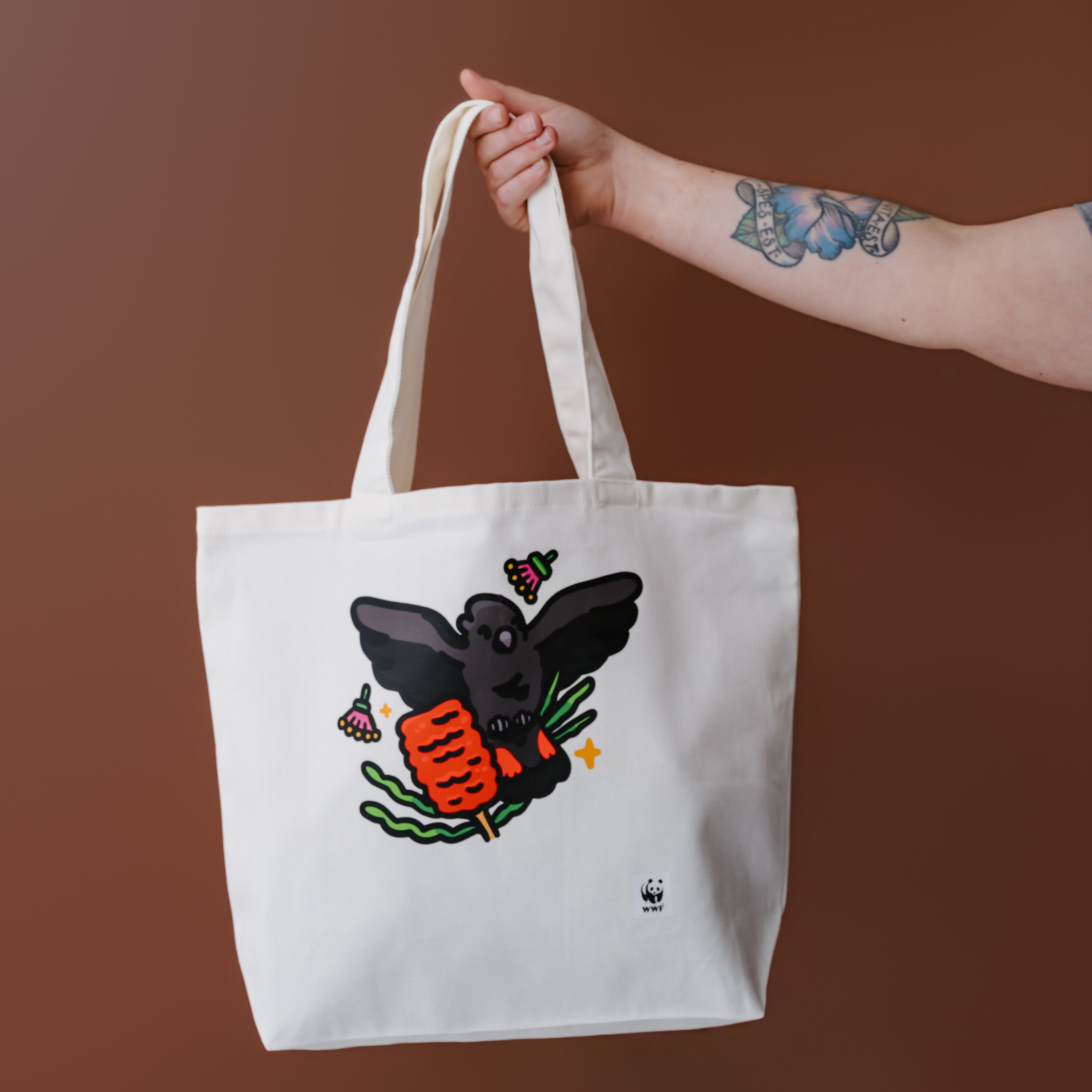 A white canvas tote bag with a graphic of a glossy black cockatoo, held by a person's tattooed arm against a solid brown background.
