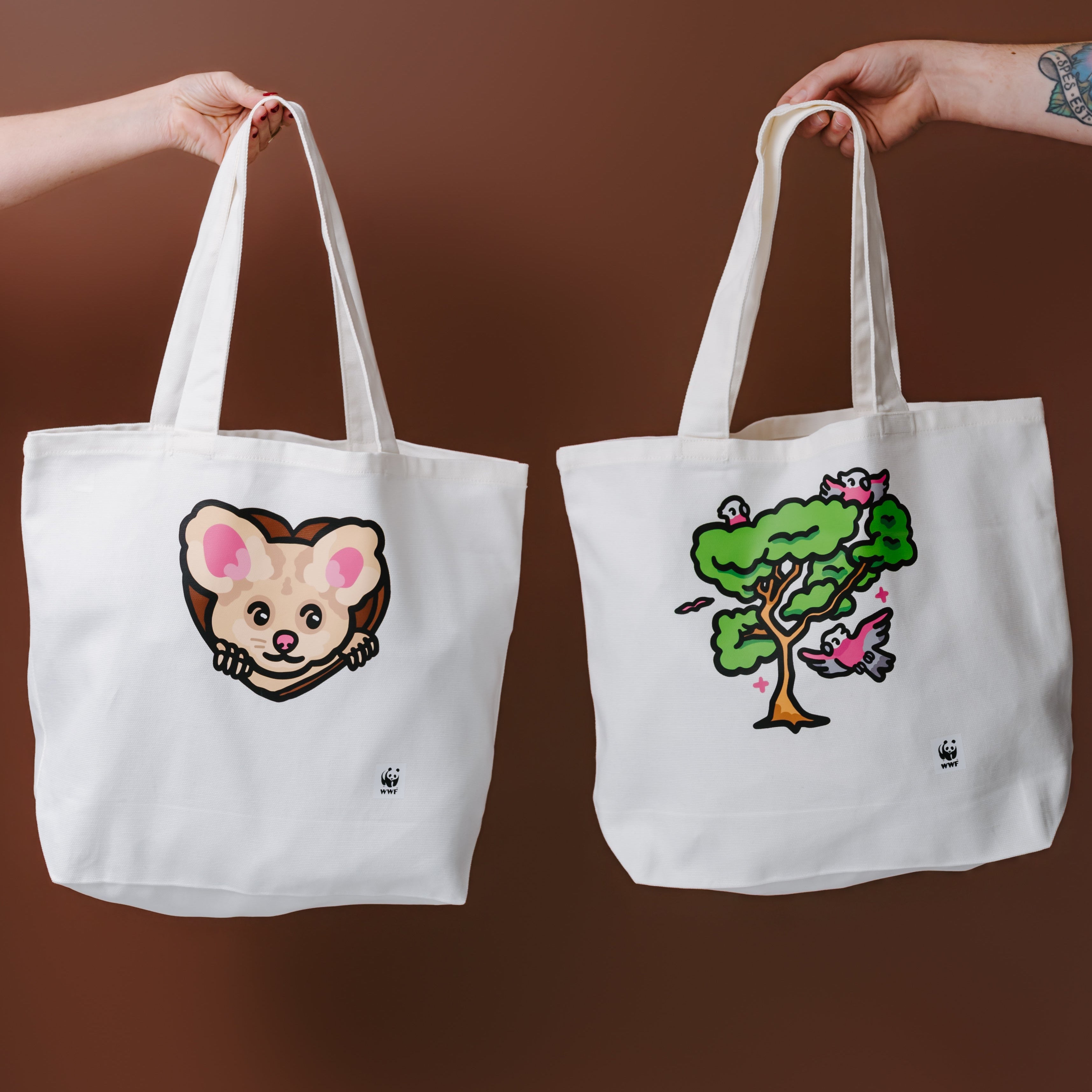 Two white canvas tote bags held up by human arms against a solid brown background. The left tote bag features a greater glider illustration, and the right tote bag features an illustrated tree with birds.