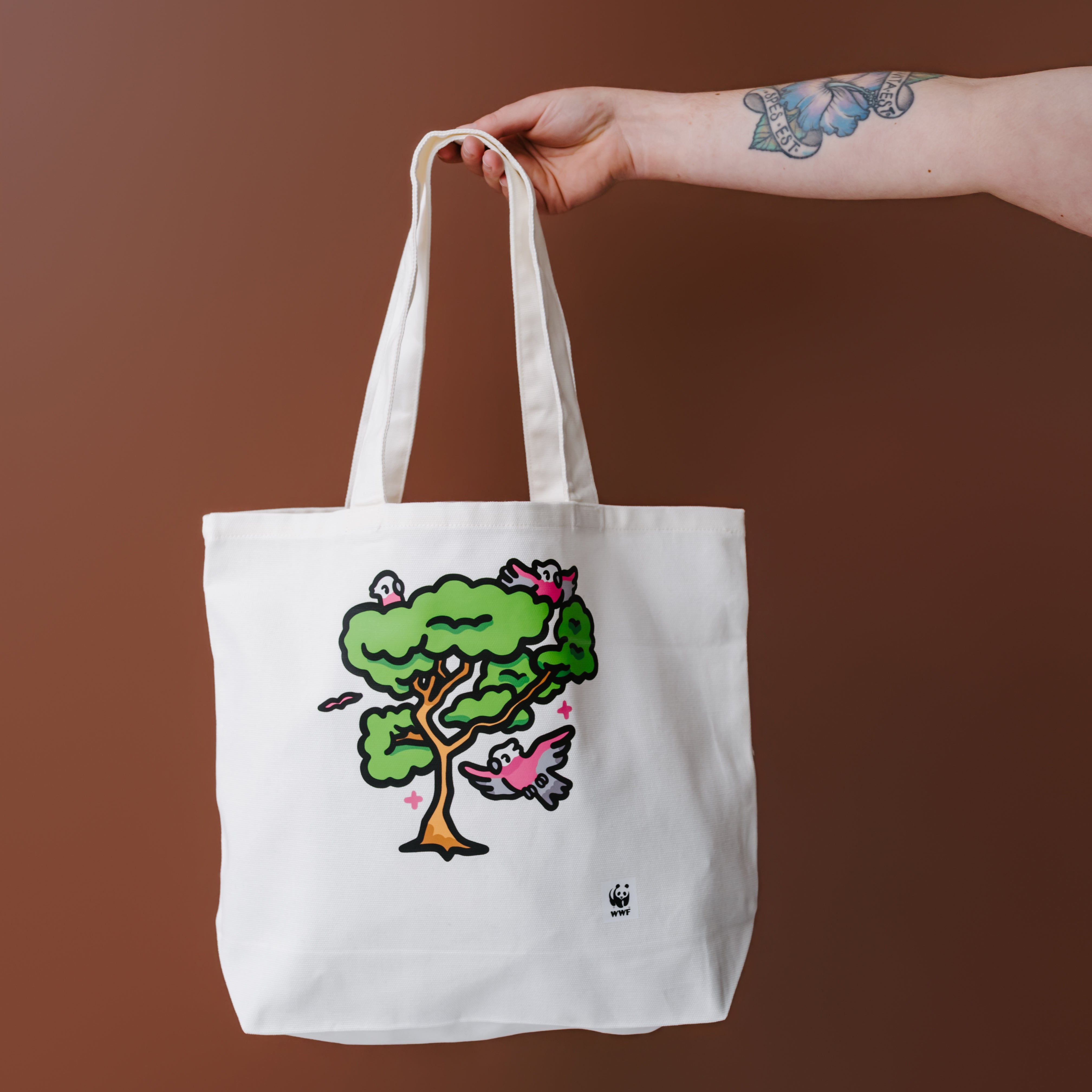 A white canvas tote bag with a colorful illustrated tree and birds graphic, held by a tattooed arm against a solid brown background.