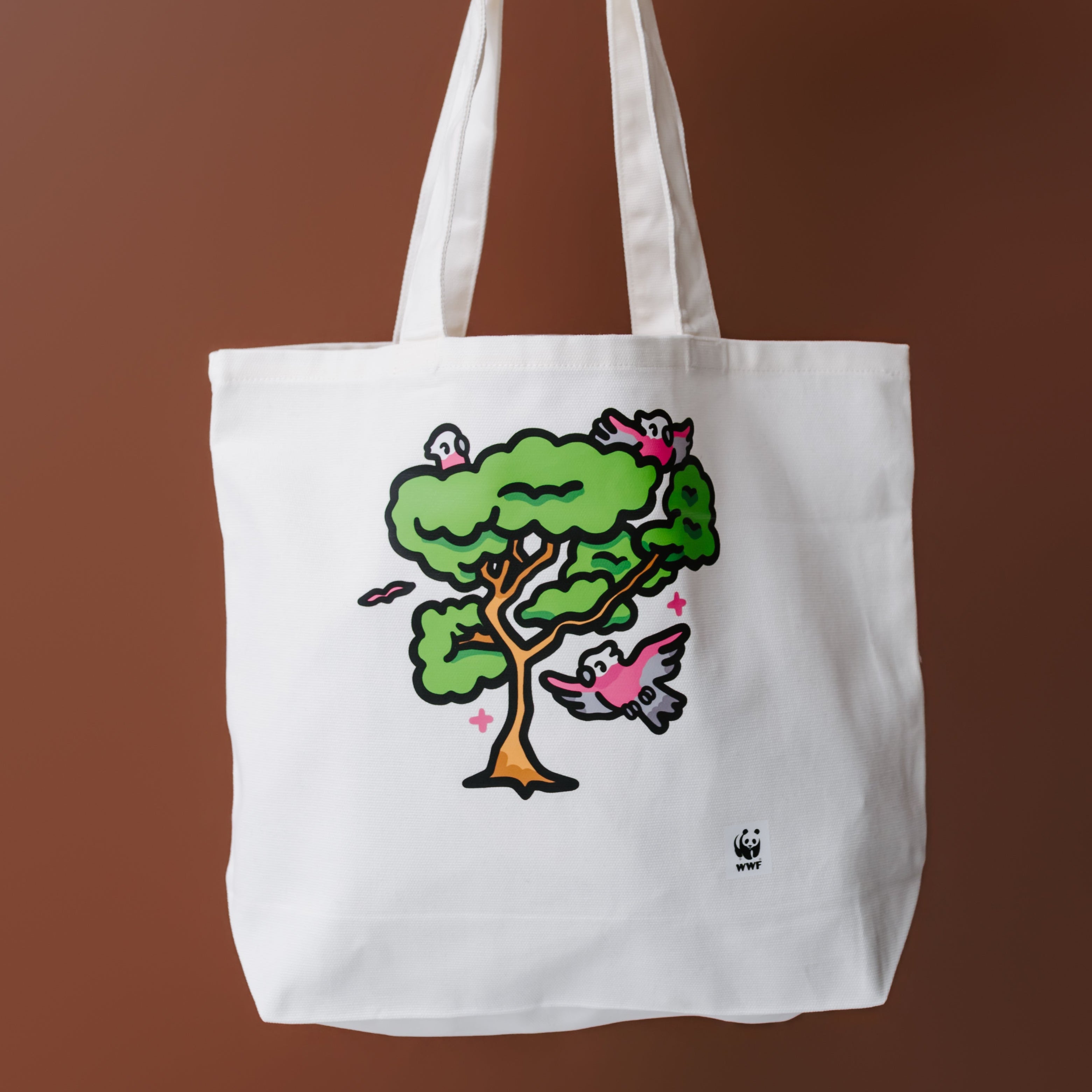 A white canvas tote bag with a colorful cartoon tree and bird graphic, held by a tattooed arm against a solid brown background.