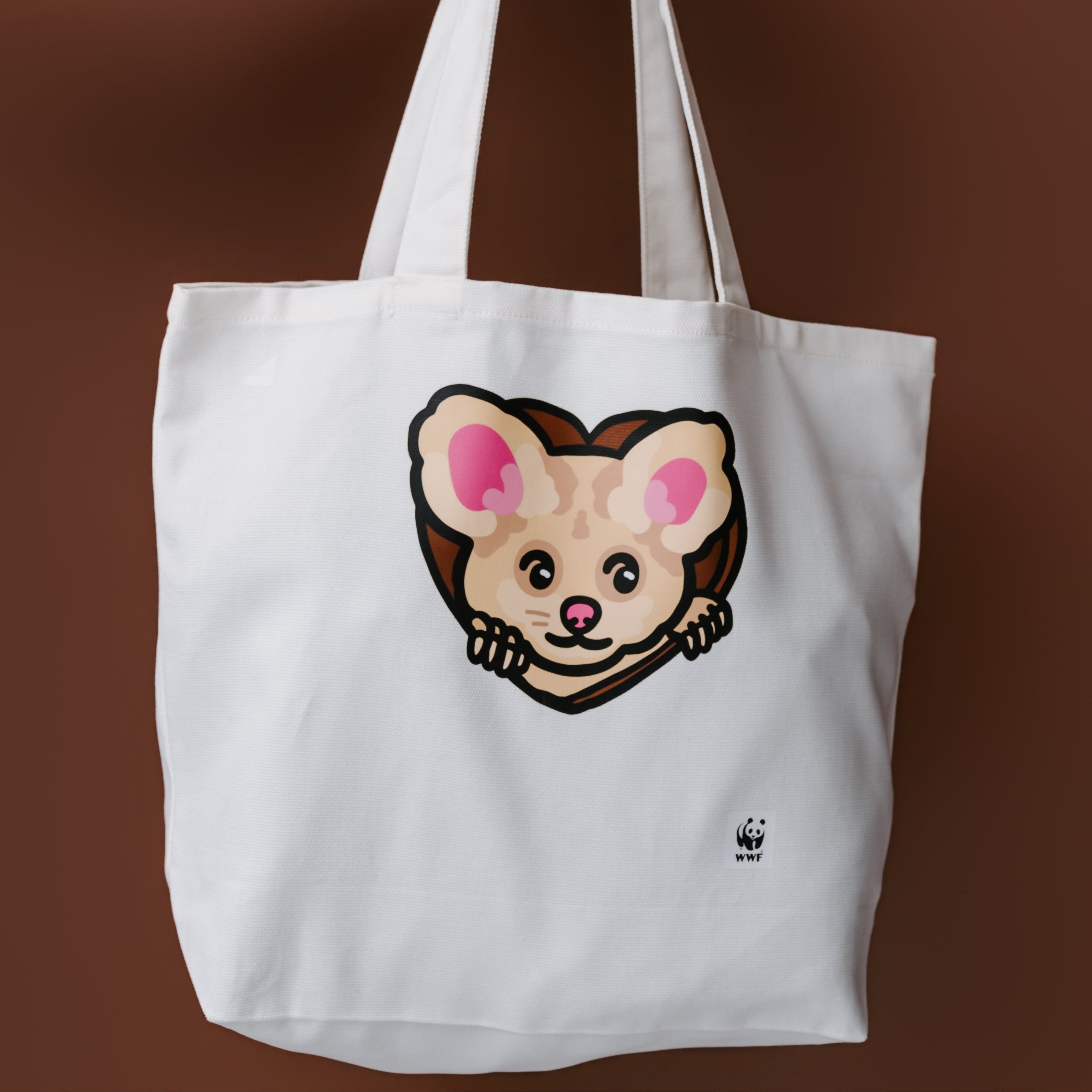 A white canvas tote bag with an illustrated greater glider graphic on the front, held by a person's arm against a solid brown background.